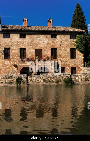 Gli edifici in pietra toscane color miele e rosata circondano il bagno di Santa Caterina del 16th secolo, una piscina rettangolare nel centro di bagno Vignoni, Toscana, Italia, piena di acqua calda di sorgente vulcanica che sgorga dal profondo sotterraneo. Le proprietà calmanti e curative dell'acqua fumante sono state godute da Etruschi, Romani, Santa Caterina da Siena, il governatore mediceo Lorenzo il magnifico, Papa Pio II, lo scrittore Charles Dickens e il Marchese de Sade. Foto Stock