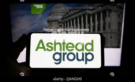 Persona che tiene uno smartphone con il logo della società di noleggio di attrezzature Ashtead Group plc sullo schermo di fronte al sito web. Mettere a fuoco sul display del telefono. Foto Stock