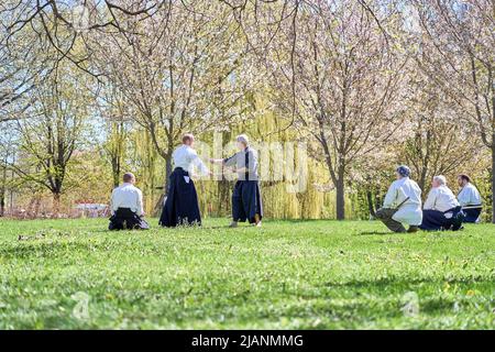 Riga, Lettonia - 8 maggio 2022: Il maestro di Aikido mostra agli allievi tecniche di combattimento con bastone di legno nel parco pubblico. sakura in fiore sullo sfondo. Foto Stock