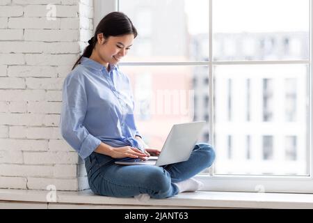 Signora asiatica positiva seduta sul davanzale, usando il laptop Foto Stock
