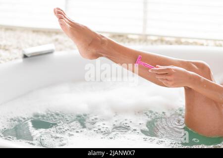 Vista ritagliata della giovane donna che prende il bagno di bolla, la rasatura della sua gamba con il rasoio, facendo la procedura domestica del centro termale, spazio di copia Foto Stock