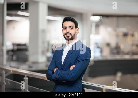 Sorridente fiducioso calma attraente giovane uomo d'affari medio-orientale con barba in vestito con le braccia incrociate Foto Stock
