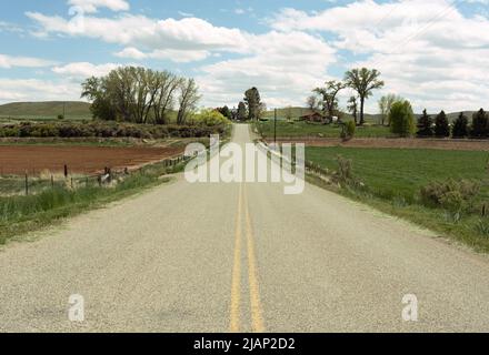 Guardando giù la linea gialla centrale di una strada a Shell, Wyoming si vede una fattoria in lontananza. Nuvole, cielo blu, alberi, pascoli sono inclusi. Foto Stock