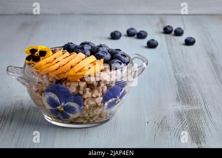 Fiocchi d'avena a grani interi in una ciotola di vetro decorata con fiori, mirtilli, fette di albicocca e semi di lino. Colazione romantica Foto Stock