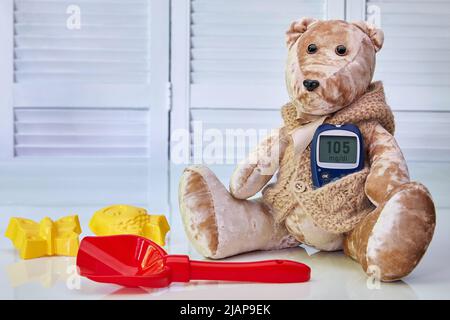 Orsacchiotto con un glucometro di sangue e giocattoli sparsi per bambini su sfondo blu. Il concetto di trattamento del diabete mellito nei bambini, hype Foto Stock