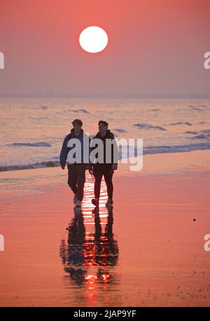 Due giovani uomini che camminano lungo la spiaggia al tramonto. Amici. Cielo rosso e riflessi nella sabbia. Bassa marea, Brighton & Hove, East Sussex, Inghilterra, Regno Unito Foto Stock