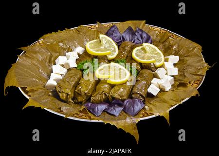 Dolma di foglie d'uva su un grande piatto circondato da foglie d'uva, basilico, prezzemolo, spicchi di limone e formaggio di cagliata. Isolare su uno sfondo nero Foto Stock