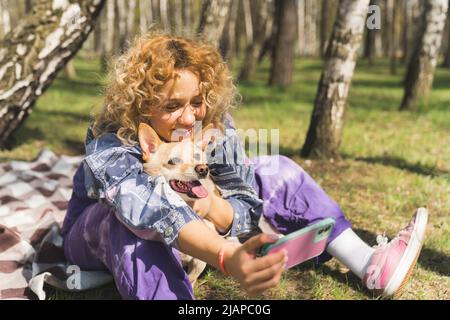 Scatto orizzontale all'aperto di una ragazza europea con capelli ricci facendo un pic-nic in un parco con il suo amico cane. Donna che prende un selfie con il suo compagno animale. Foto di alta qualità Foto Stock