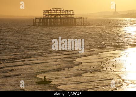 Forme di uomo con kayak sulla costa a bassa marea, Brighton & Hove, East Sussex, Inghilterra, Regno Unito. Resti del dilapidato West Pier oltre. Gabbiani sulla riva. Foto Stock