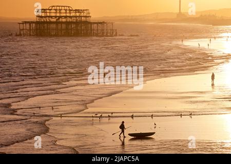 Forme di uomo con un kayak sulla spiaggia a bassa marea, Brighton & Hove, East Sussex, Inghilterra, Regno Unito. Fotografato dal Palace Pier con i resti del West Pier, e Shoreham Power Station in lontananza. A piedi dal mare Foto Stock