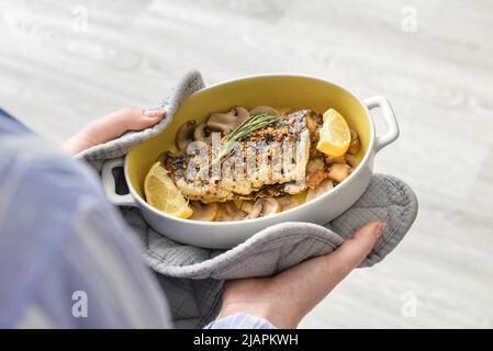 Donna che tiene piatto da forno con gustoso pesce branzino in cucina Foto Stock