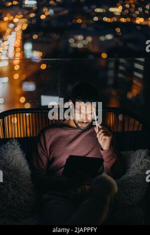 Giovane afroamericano seduto sul balcone con vista sulla città e con un tablet di notte Foto Stock