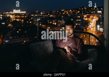 Giovane afroamericano seduto sul balcone con vista sulla città e con un tablet di notte Foto Stock