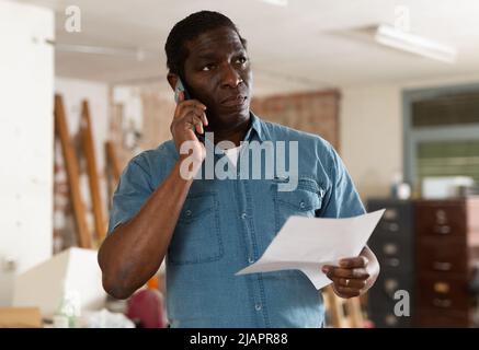 Perplesso africano americano con carte che parlano al telefono durante la ristrutturazione della casa Foto Stock