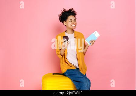 Gioiosa ragazza afroamericana ricci capelli, ha il passaporto con i biglietti di viaggio e smart phone, siediti su valigia gialla su sfondo rosa isolato, guardando al lato, sogni di vacanza, sorridente Foto Stock