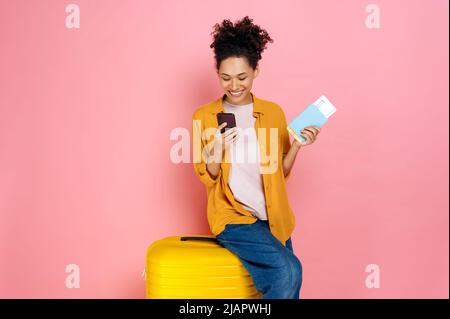 Felice bella, ragazza africana americana ricci capelli, indossare abiti casual, tenendo passaporto con biglietti di viaggio, seduto su valigia gialla su sfondo rosa isolato, utilizza il suo smartphone, sorridente Foto Stock