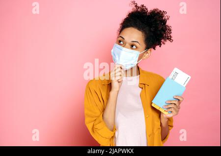 Ragazza frulla pensiva afroamericana ricci con maschera medica sul viso, con passaporto e biglietti di viaggio, in piedi su sfondo rosa isolato, guardando lateralmente, sognando di vacanza Foto Stock