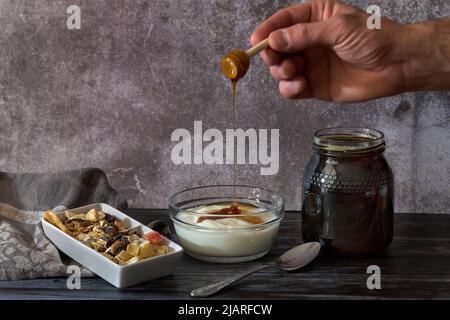 Una mano di una persona che versa il miele naturale sopra lo yogurt fatto in casa fresco e le noci in un contenitore Foto Stock