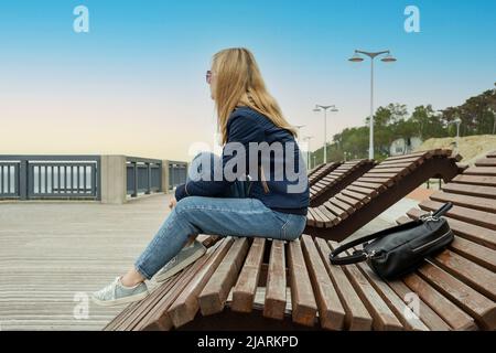 Una giovane donna bionda solitaria su un argine vuoto della città riposa seduta su un lettino da sole in una serata di primavera o autunno e guarda in lontananza. Il Foto Stock
