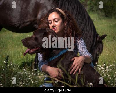 Vista orizzontale della femmina con il suo labrador Retriever cioccolato in montagna in prato primaverile e cavallo nero sullo sfondo. Foto Stock