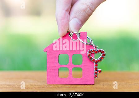 Mano che tiene uno scheletro chiave a forma di cuore decorato con rhinestones rossi e una casa rosa piatta a forma di portachiavi. Trasferirsi in una casa di campagna. Foto Stock