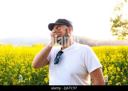 Uomo che tiene la mano in faccia con stupore e imbarazzo. Campo di canola e allergie. Foto Stock