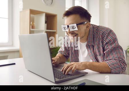 Strano uomo assonnato stanco con divertente carta adesivo occhi che lavorano sul computer portatile Foto Stock