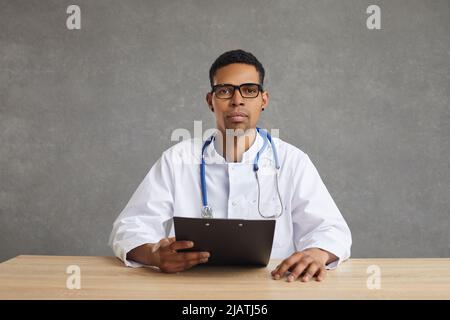 Serio medico afroamericano che guarda la macchina fotografica seduta allo studio di scatto della scrivania Foto Stock