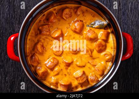 nakkikastike, salsicce finlandesi, bagna in salsa di pomodoro cremosa in pentola rossa su tavola di legno scuro, vista orizzontale dall'alto, posa piatta, primo piano Foto Stock