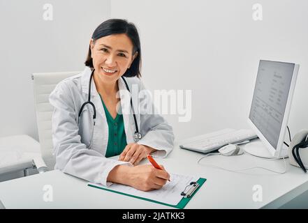 Ritratto di medico generico donna che indossa il cappotto del medico seduto nel suo ufficio sul posto di lavoro presso la clinica medica. consulto medico Foto Stock