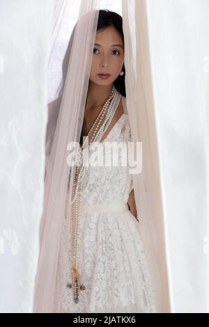 Una bella donna asiatica che indossa un abito in pizzo bianco e posa tra tessuto velato scorrevole Foto Stock