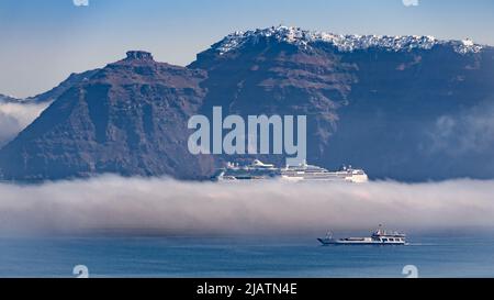 Una nuvola del mattino presto sdraiata sull'acqua della caldera di Santorini oscura parzialmente una nave da crociera che viene passata da un traghetto che si muove alla luce del sole Foto Stock