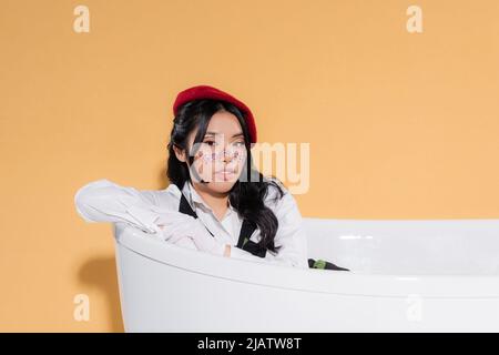 Elegante donna asiatica in guanti e berretto seduta in vasca da bagno su sfondo arancione Foto Stock