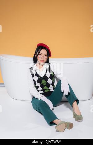 Donna asiatica alla moda in beretto e guanti seduti vicino vasca da bagno su sfondo arancione Foto Stock