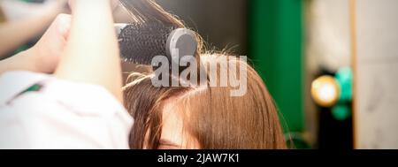 Un parrucchiere asciuga capelli lunghi e marroni con un asciugacapelli e una spazzola rotonda in un salone di bellezza Foto Stock