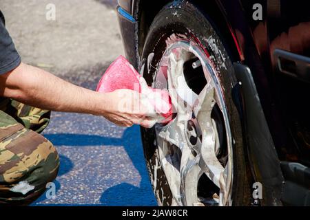 L'uomo lava la ruota dell'auto. La mano maschio contiene una spugna rosa con schiuma saponata per la pulizia. Autolavaggio in cortile. Foto Stock