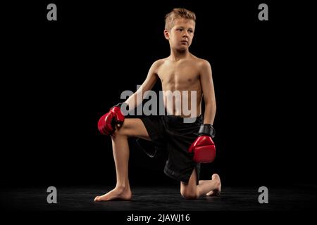 Scatto dello studio del ragazzo di preschool, capretto carino nei colpi e nei guanti di sport che posano su sfondo nero. Sport, educazione, azione, motion concept. Foto Stock