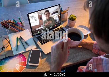 Un uomo d'affari caucasico che ha un caffè durante una videoconferenza con un collega maschile asiatico Foto Stock