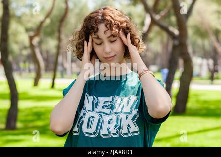 Giovane donna rossa stressata che indossa una tee verde in piedi sul parco della città, all'aperto tenendo la testa con le mani cercando di ricordare qualcosa o avere la testa Foto Stock