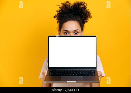 Stupito bruna ragazza afroamericana entusiasta che si alza da dietro il laptop, guarda sorpreso la fotocamera, si erge su sfondo arancione isolato, tiene un portatile aperto con bianco bianco bianco mock-up schermo Foto Stock