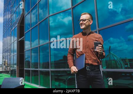 Un giovane uomo attento e bello in bicchieri con un portatile e un bicchiere di caffè da andare, si trova sulla terrazza di un ufficio. Giovane sviluppatore intelligente Foto Stock