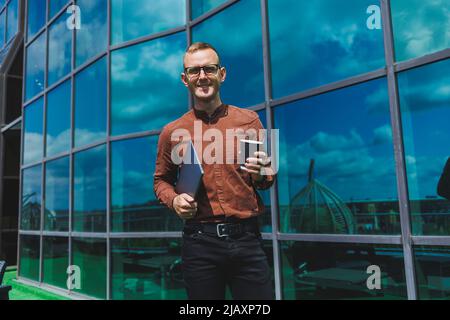 Un giovane uomo attento e bello in bicchieri con un portatile e un bicchiere di caffè da andare, si trova sulla terrazza di un ufficio. Giovane sviluppatore intelligente Foto Stock