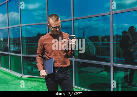 Un giovane uomo attento e bello in bicchieri con un portatile e un bicchiere di caffè da andare, si trova sulla terrazza di un ufficio. Giovane sviluppatore intelligente Foto Stock