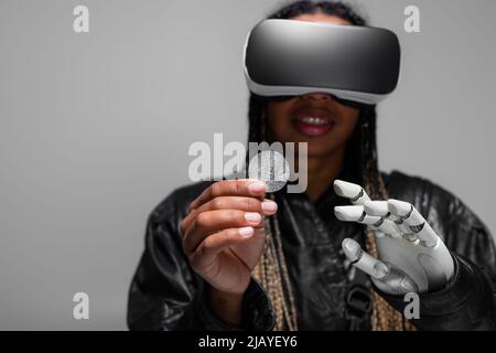 KYIV, UCRAINA - 28 APRILE 2022: Donna afro-americana offuscata con visore vr e bitcoin robotico in mano isolato su grigio Foto Stock