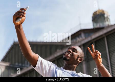 Da sotto di felice giovane afroamericano maschio turista. Sta mostrando due dita di gesto e sorridendo luminoso mentre prende selfie su smartphone contro la chiesa in giorno di sole Foto Stock