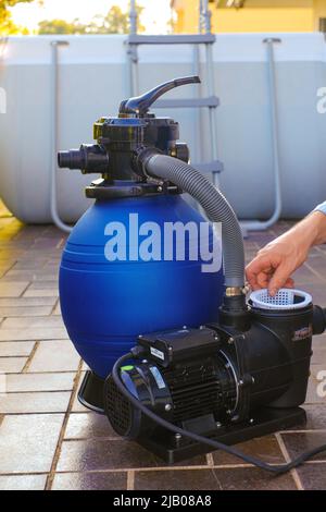 Attrezzatura per la pulizia della piscina. Filtro dell'acqua nelle mani di un uomo sul terrazzo vicino alla casa.l'uomo sposta i tubi al filtro nel primo piano della piscina. Foto Stock