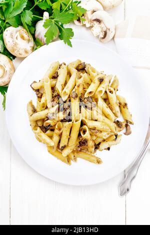 Penne pasta con funghi selvatici in un piatto, asciugamano, prezzemolo e forchetta su un fondo di legno chiaro dall'alto Foto Stock