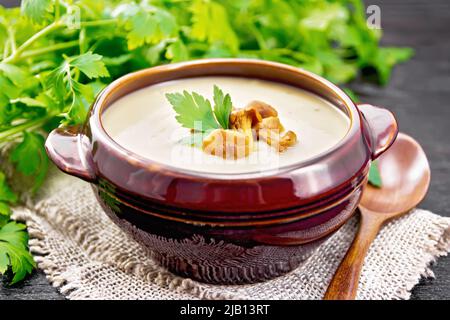 Zuppa di purea di craterelle, patate, cipolle e panna in una ciotola di argilla su burlap, prezzemolo e un cucchiaio su sfondo nero tavola di legno Foto Stock