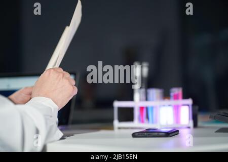 Le mani del medico che tiene il documento sullo sfondo di un set di utensili da laboratorio per la ricerca medica o scientifica. Un bicchiere con prodotti chimici. Foto di alta qualità Foto Stock