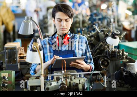 Serio concentrato giovane impiegato di fabbrica femminile occupato con i capelli corti neri che indossano le protezioni dell'orecchio sul collo usando il tablet mentre sincronizza con Foto Stock